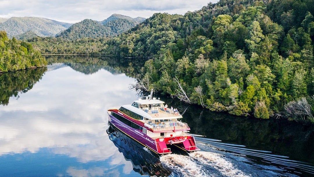 Red cruise boat on Gordon River, surrounded by lush forest and mountains, World Heritage site.