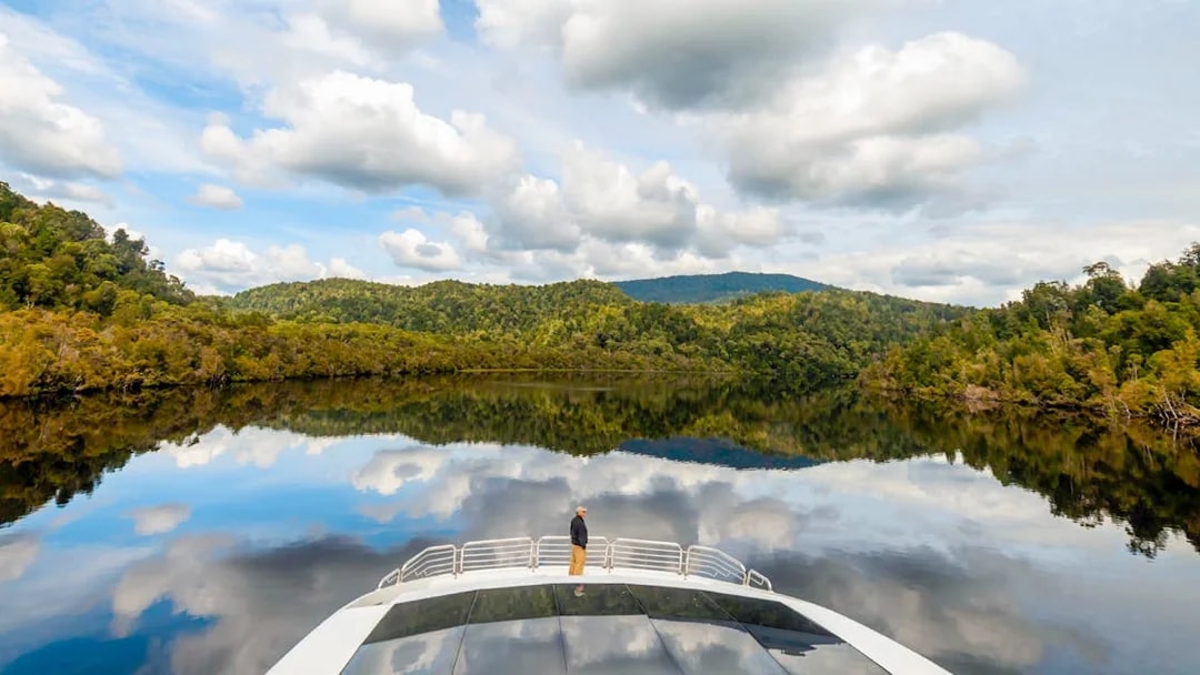Man on cruise boat on Gordon River, surrounded by lush forest and calm water reflections.