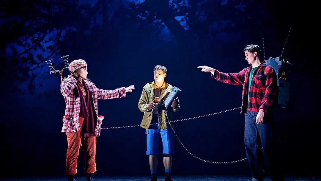Three actors on stage in a scene from "Stranger Things: The First Shadow," with one holding a device.