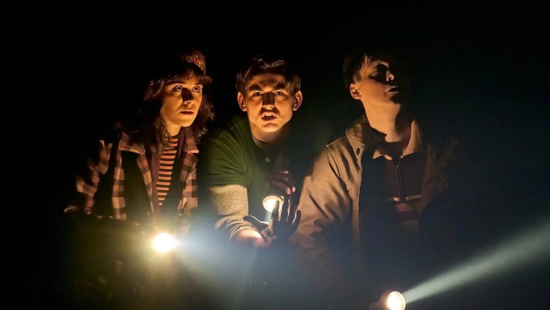 Three people holding flashlights in a dark setting, related to Stranger Things: The First Shadow.
