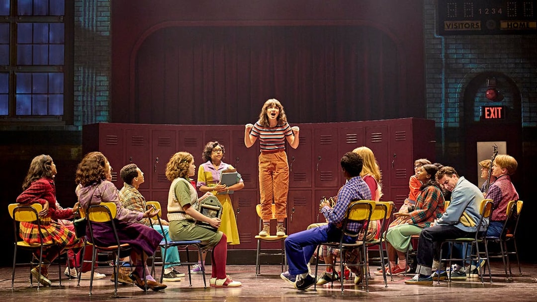 Group of students in a school setting, one standing on a chair, from Stranger Things: The First Shadow.