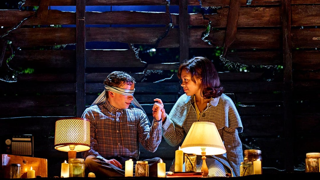 Two people in a dimly lit room with candles, one blindfolded, from "Stranger Things: The First Shadow.