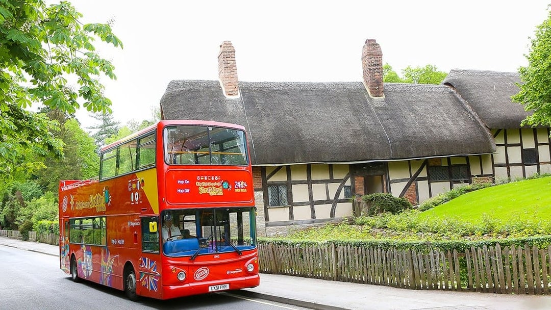 Red double-decker HOHO tour bus in Stratford passing a thatched cottage.