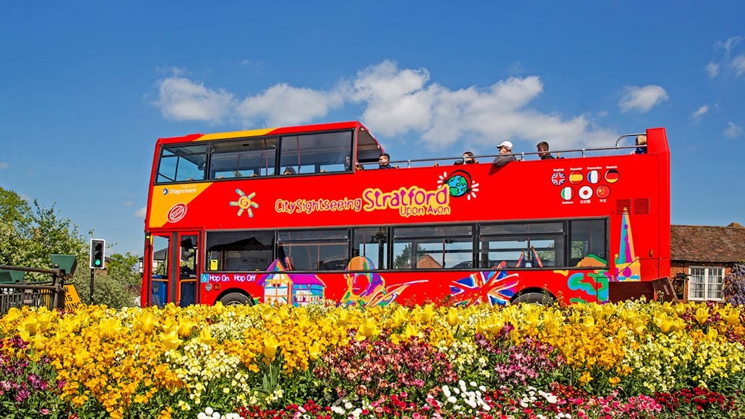 Red double-decker bus on City Sightseeing tour in Stratford-upon-Avon, passing colorful flower beds.