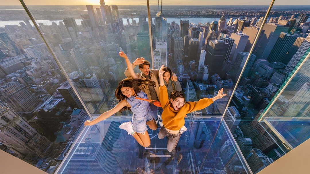 Four people posing on a glass platform above a city skyline at sunset.