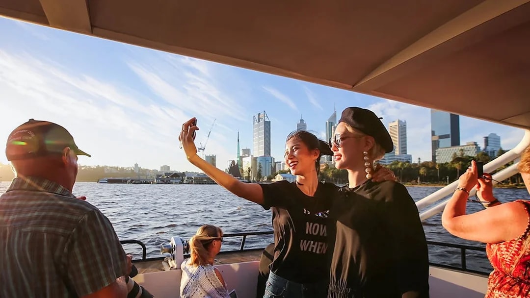 Tourists taking a selfie on a boat during sunset with city skyline in the background.