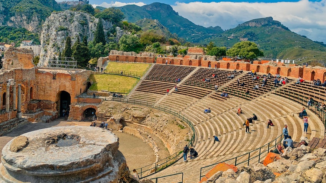 Taormina Ancient Theatre with visitors seated, surrounded by scenic hills.