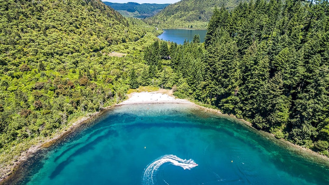 Aerial view of Lake Tarawera with lush green forest and a boat creating a wake in the water.