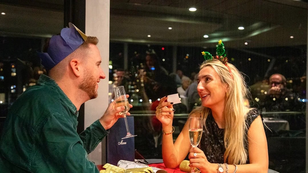 Guests enjoying Christmas dinner on Thames River cruise with festive hats and champagne.