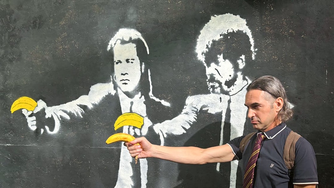 Man posing with Banksy mural of figures holding bananas at The Banksy Museum.