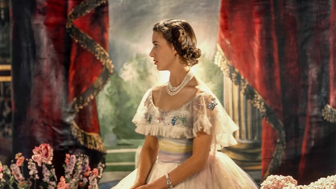 A woman in an elegant gown with pearls, seated in a regal setting with red drapes and flowers.