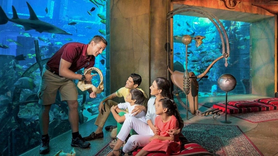 Family observing marine life at The Lost Chambers Aquarium, Dubai, with a guide showing a shark jaw.