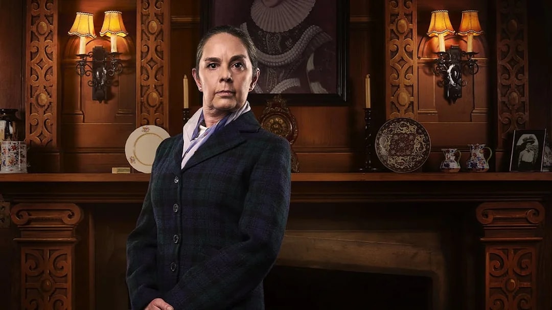 Person in period costume standing in front of ornate wooden fireplace, related to The Mousetrap.