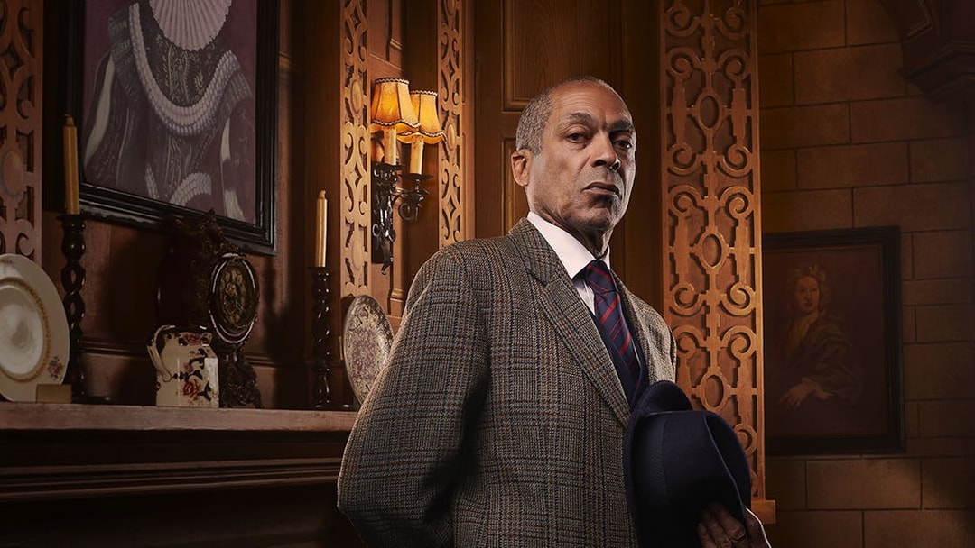 Man in a suit holding a hat, standing in an ornate room with vintage decor, related to The Mousetrap.