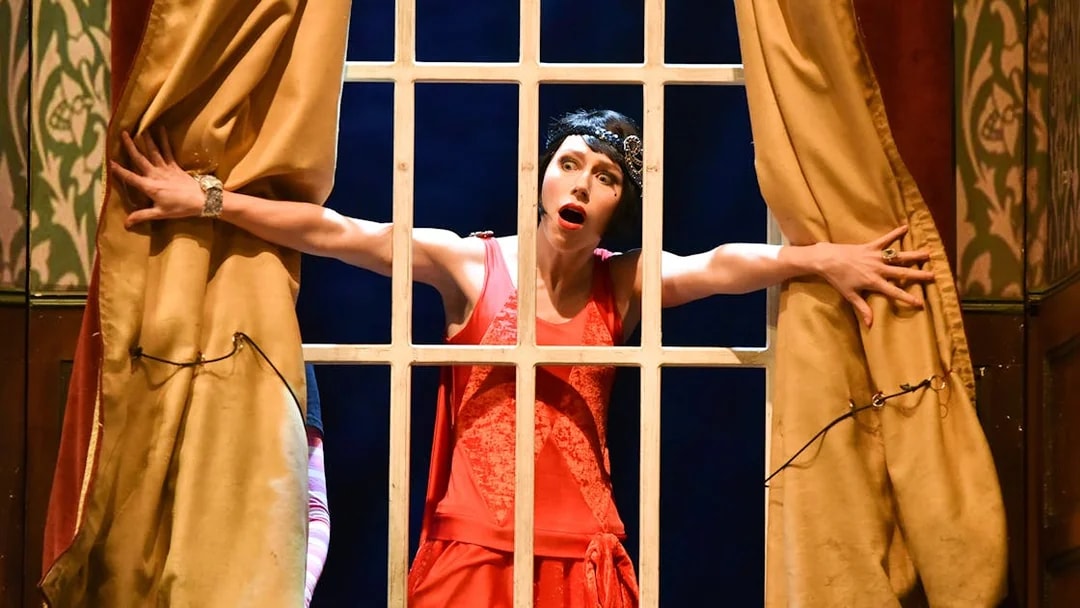 Performer in red dress on stage during "The Play That Goes Wrong" scene.