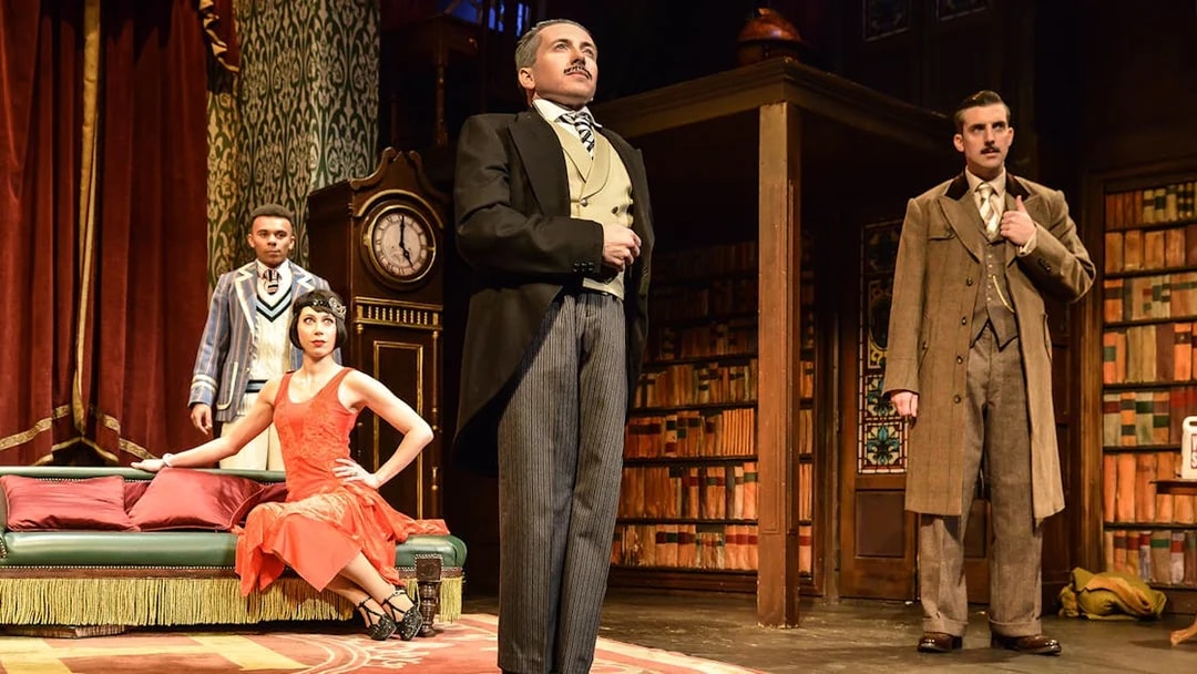 Actors performing a scene from "The Play That Goes Wrong" in a vintage library setting.