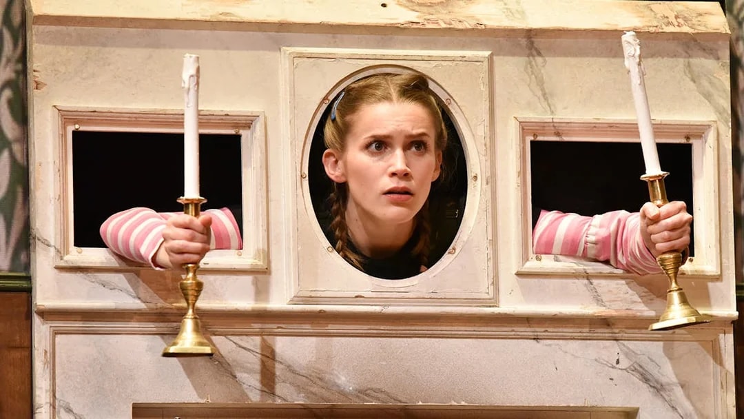 Actor peering through a wall with candlesticks in "The Play That Goes Wrong" scene.