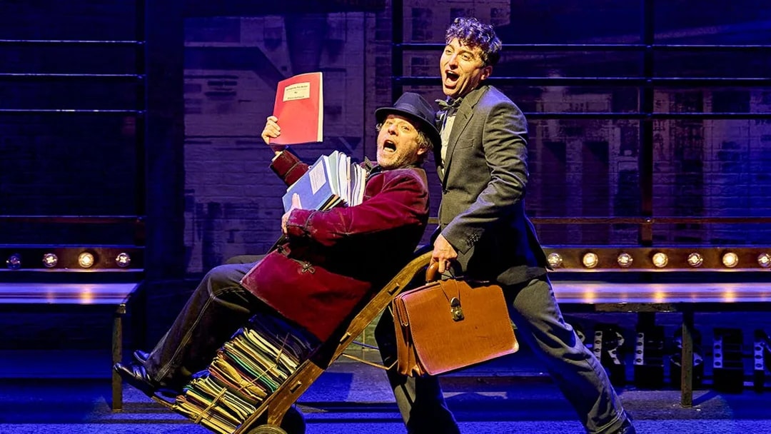 Two actors perform a comedic scene from "The Producers" on stage, one pushing a cart of scripts.