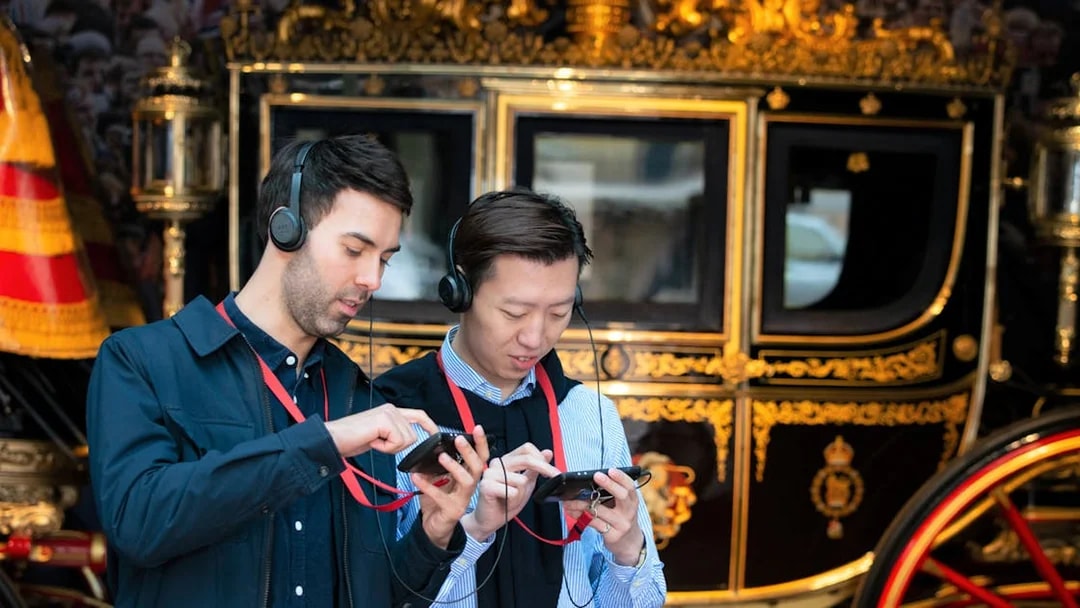 Visitors using audio guides at Buckingham Palace with a royal carriage in the background.