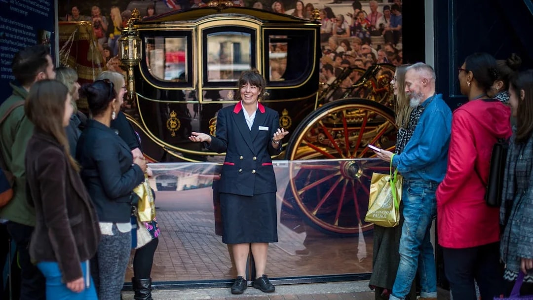 Warden guiding family visitors at The Royal Mews, London, with a historic carriage in the background.