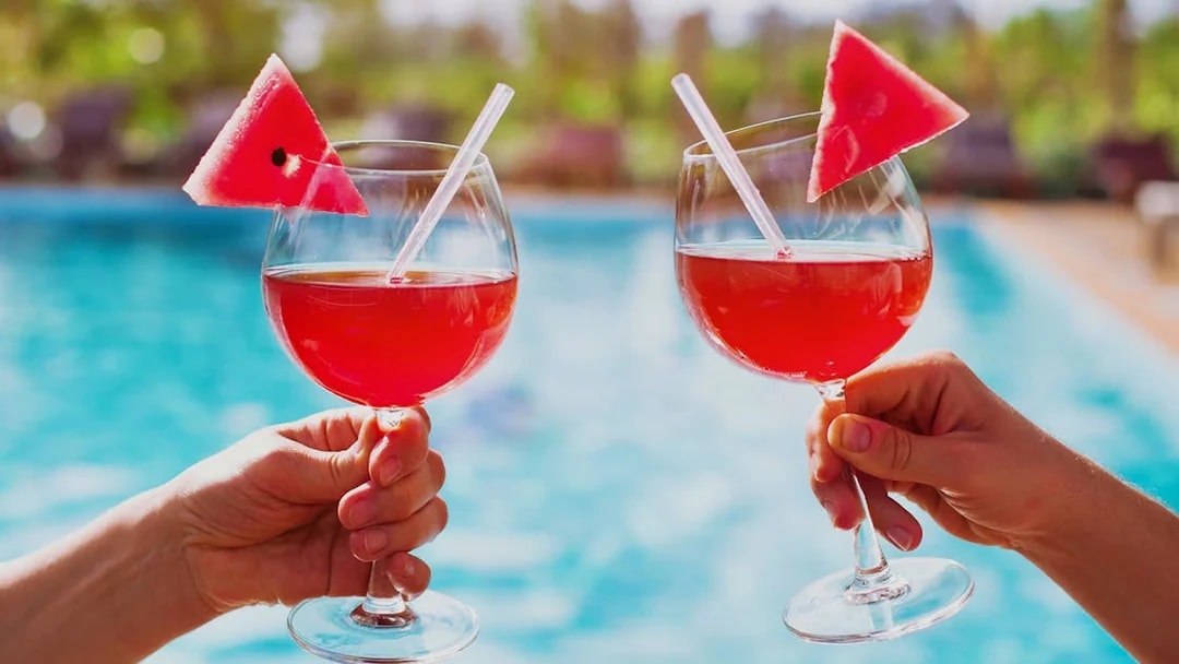 Two hands holding glasses of red drinks with watermelon slices by a pool.