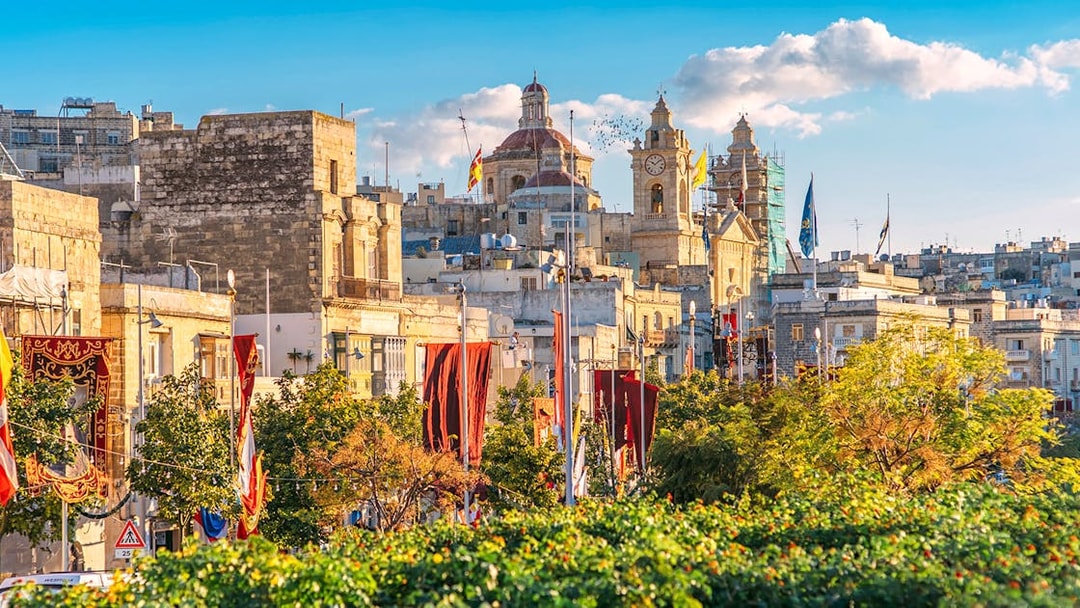 Historic buildings and clock tower in the Three Fortified Cities, Malta, with colorful banners.