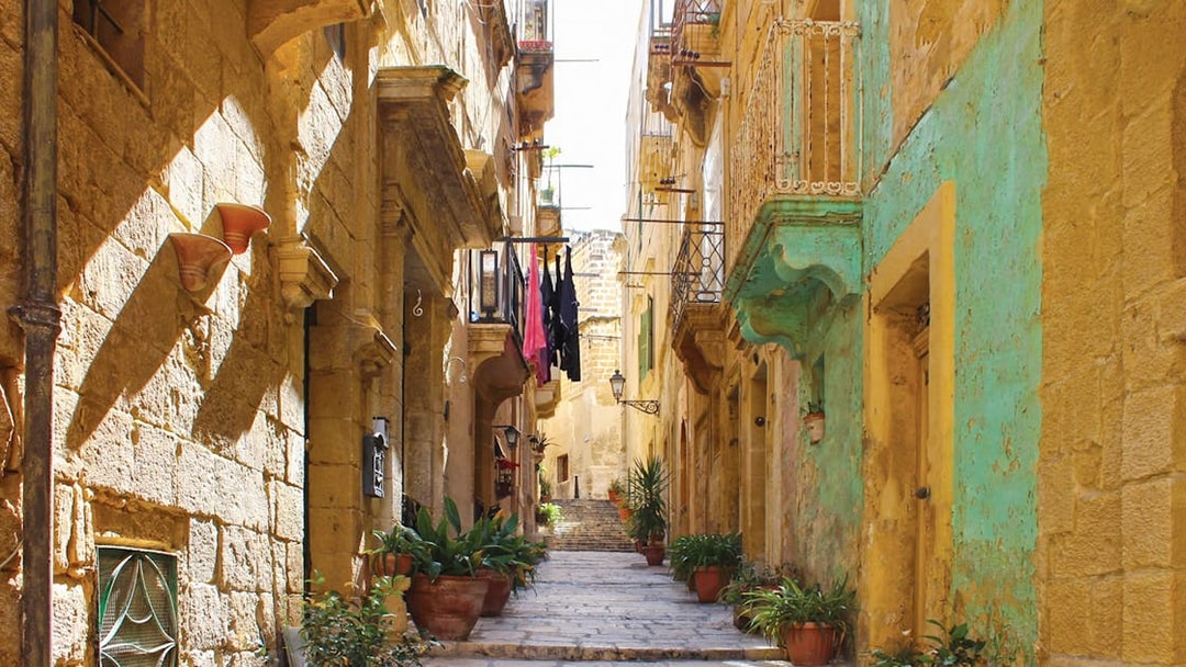 Narrow street in one of the Three Fortified Cities, lined with historic stone buildings and potted plants.