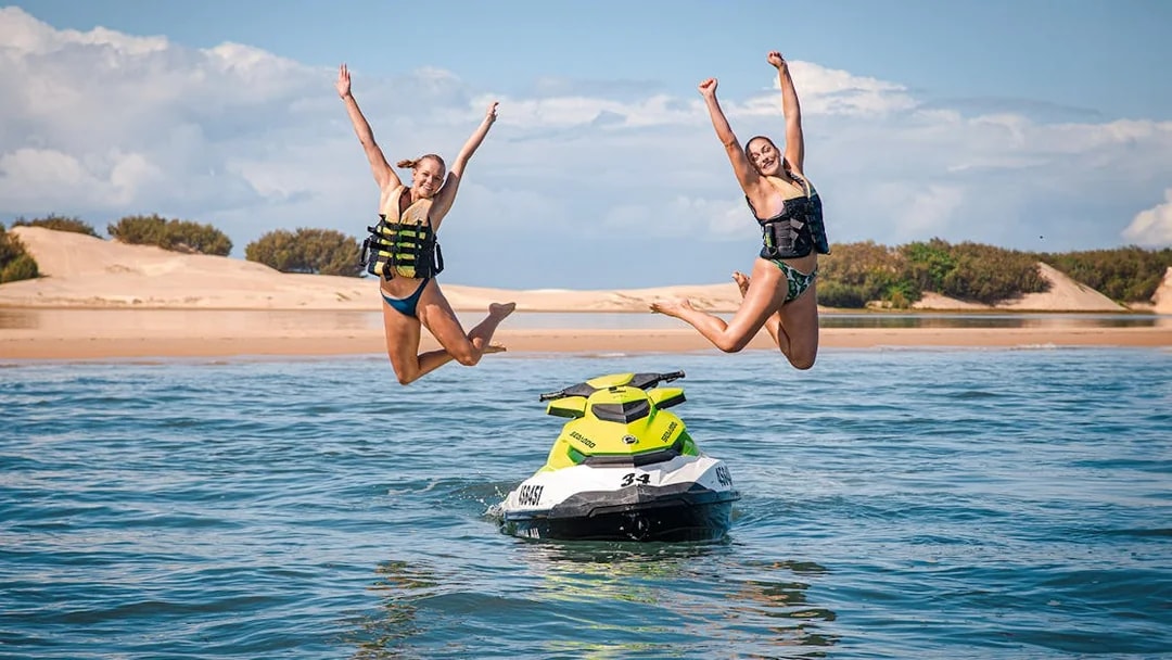Jet ski with two people jumping into the water, beach in the background.
