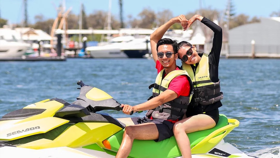 Couple enjoying a jet ski ride on a sunny day near a marina.