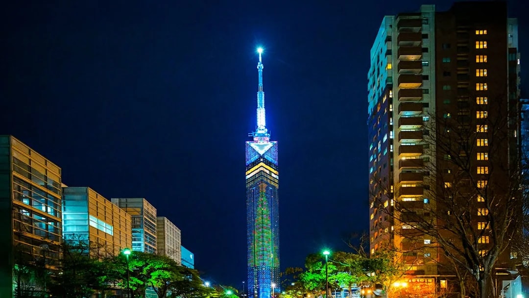 Fukuoka Tower illuminated at night, surrounded by city buildings.
