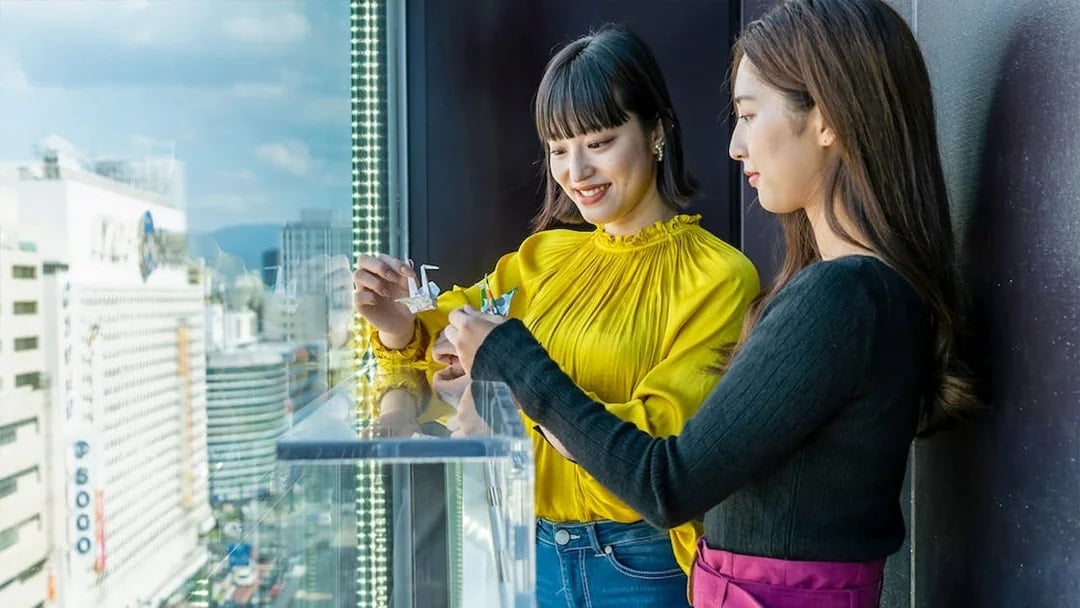 Visitors folding origami cranes at Hiroshima Orizuru Tower with city view.