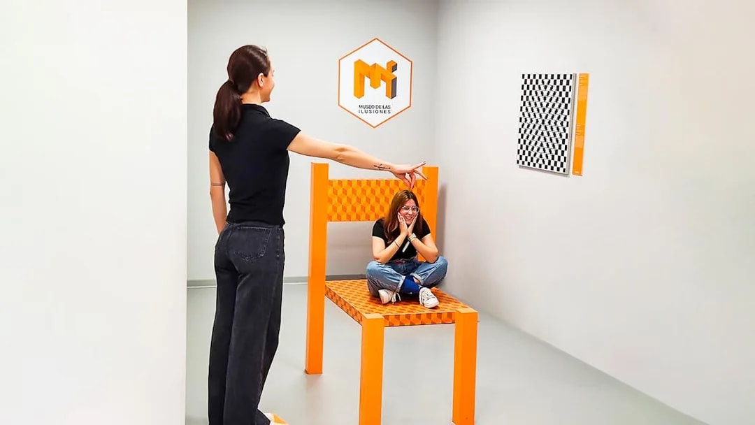 Person pointing at another sitting on oversized chair in perspective room, Museum of Illusions Seville.