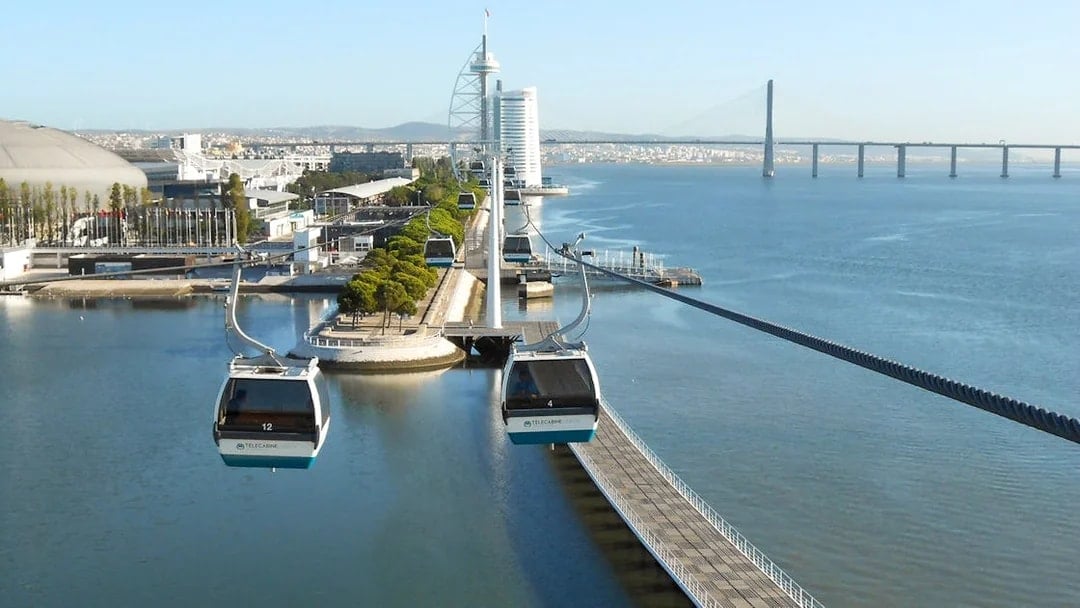Telecabine cable cars over Tagus River in Lisbon with cityscape and bridge in view.