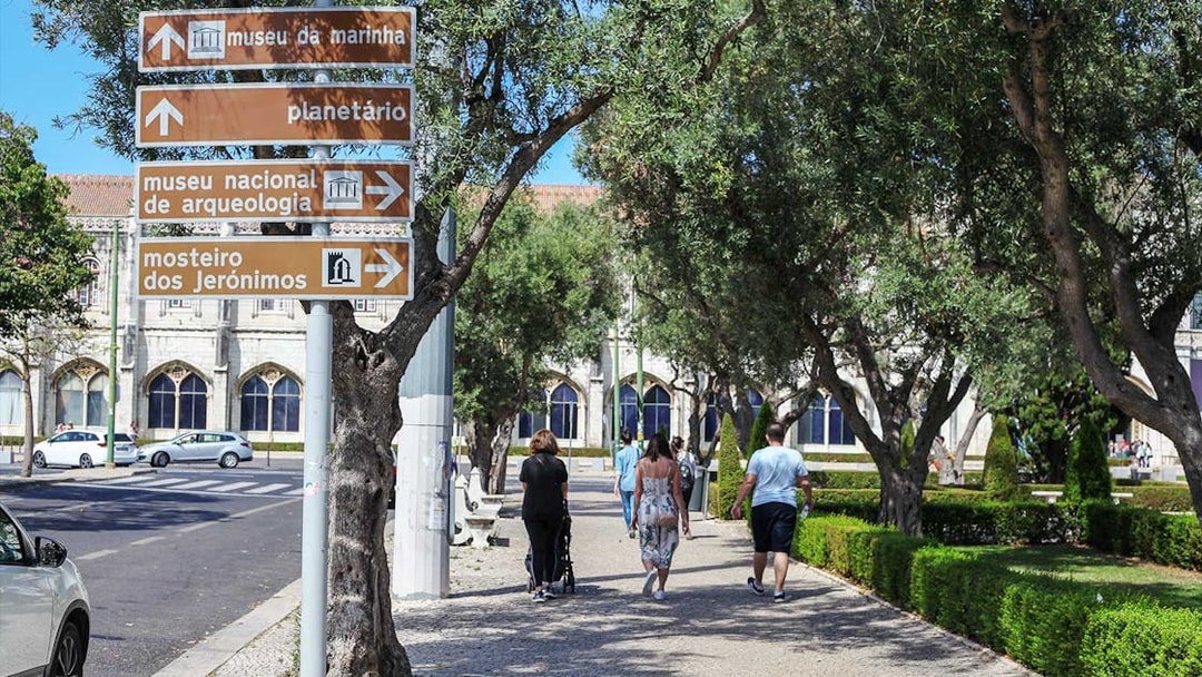 Signage for Jerónimos Monastery and Navy Planetarium in Lisbon, with people walking nearby.