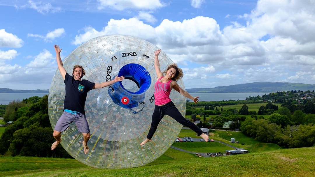 People jumping in front of a Zorb inflatable ball on a grassy hill with scenic landscape.
