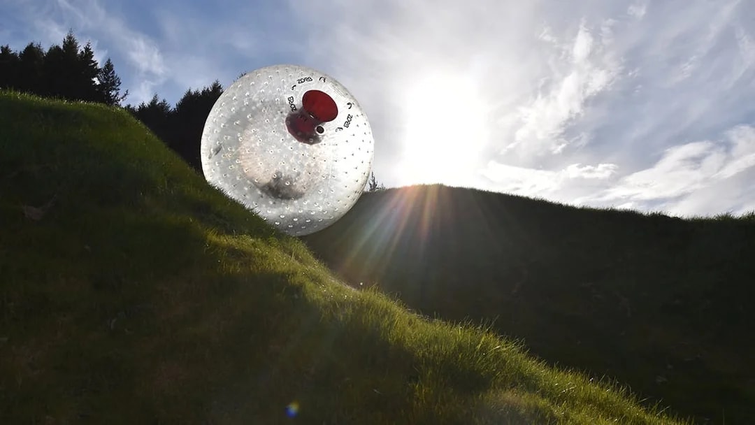 Zorb ball rolling down a grassy hill under a bright sky.