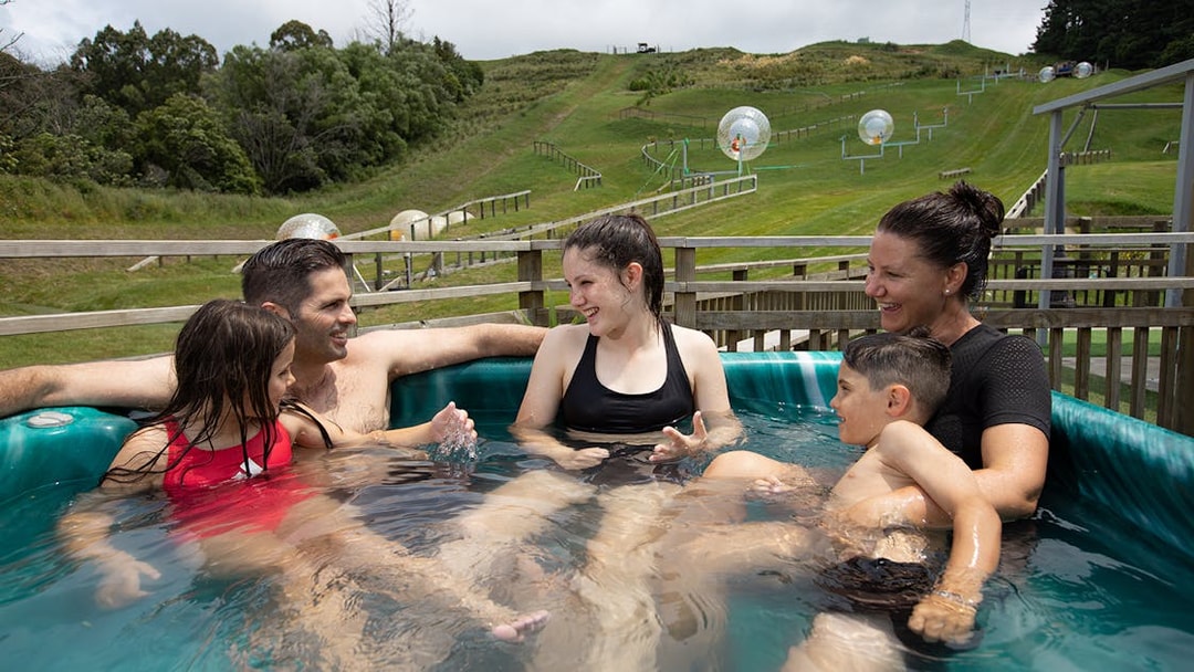Family relaxing in a hot tub with Zorb inflatable balls on a hillside in the background.
