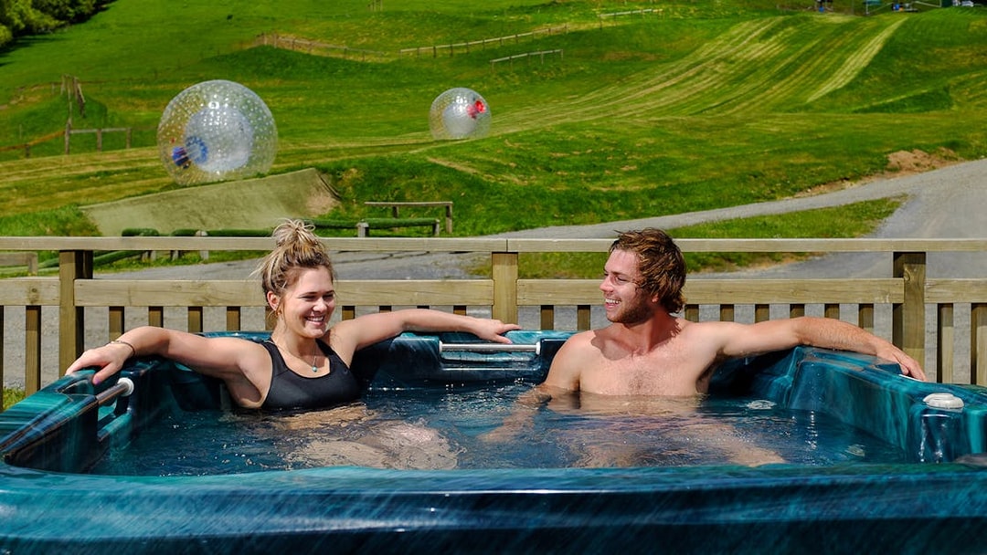 Couple relaxing in a hot tub with Zorb inflatable balls on a grassy hill in the background.