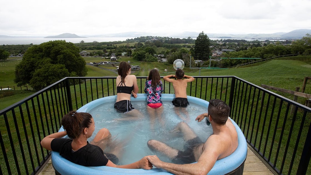 Family in hot tub overlooking landscape with Zorb ball ride in the distance, Rotorua, New Zealand.