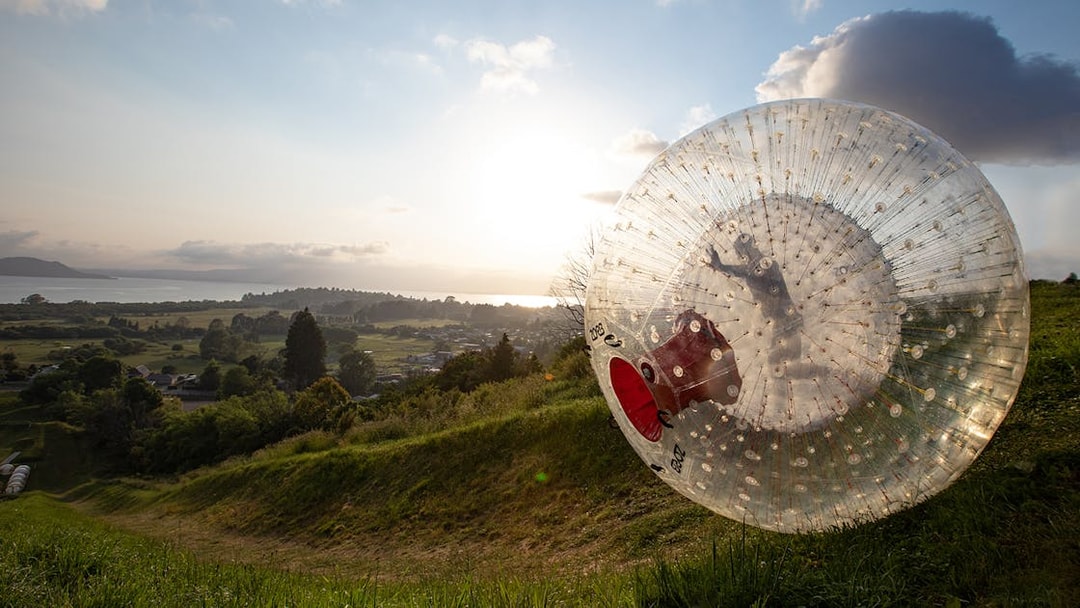 Person rolling downhill in a giant inflatable Zorb ball at sunset, overlooking a scenic landscape.