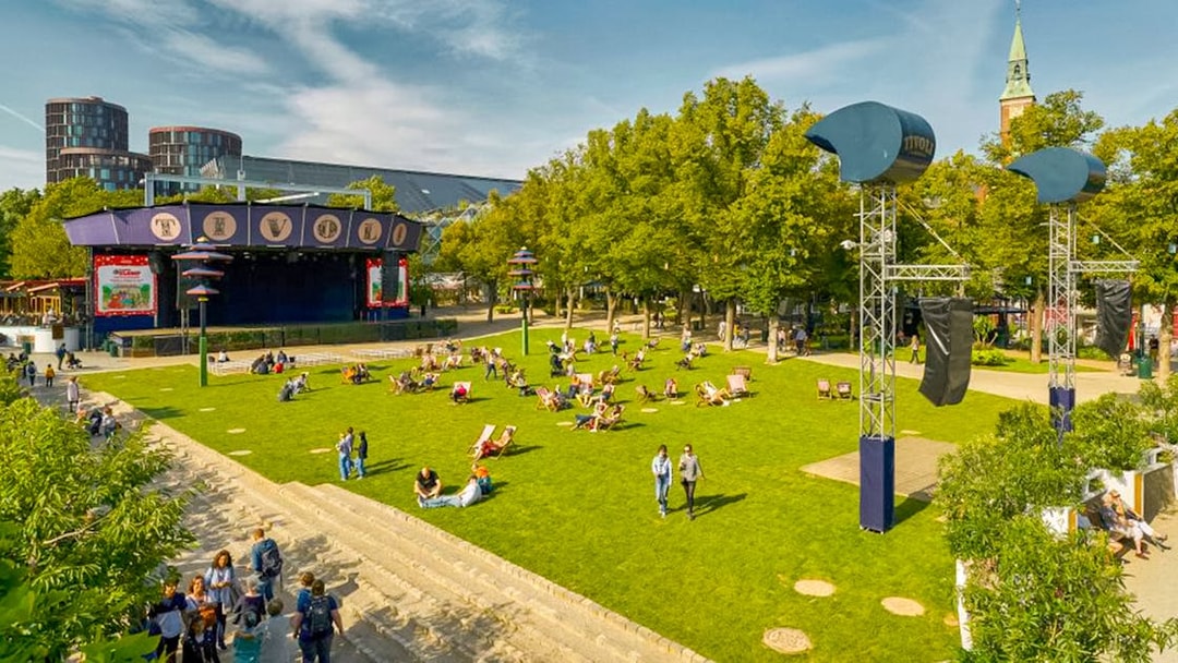 Visitors relaxing on the lawn at Tivoli Gardens, Copenhagen, with nearby attractions and rides.