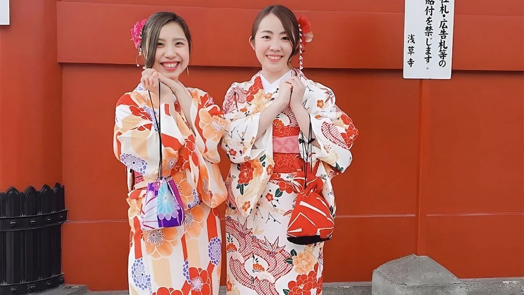Two people in colorful kimonos smiling in front of a red wall in Asakusa, Tokyo.