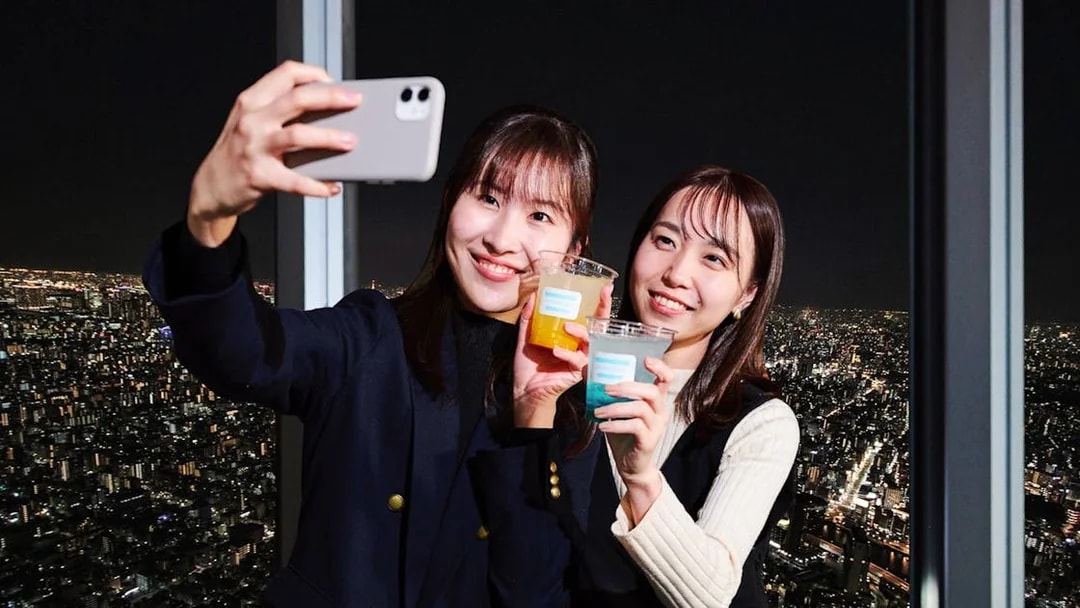 Tourists taking a selfie with drinks on Tembo Deck, Tokyo at night.