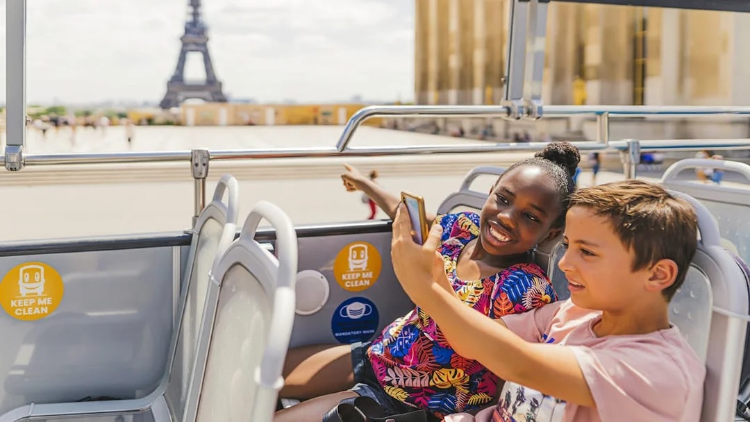 Children on a double-decker bus enjoying an audio-guided tour with the Eiffel Tower in the background.