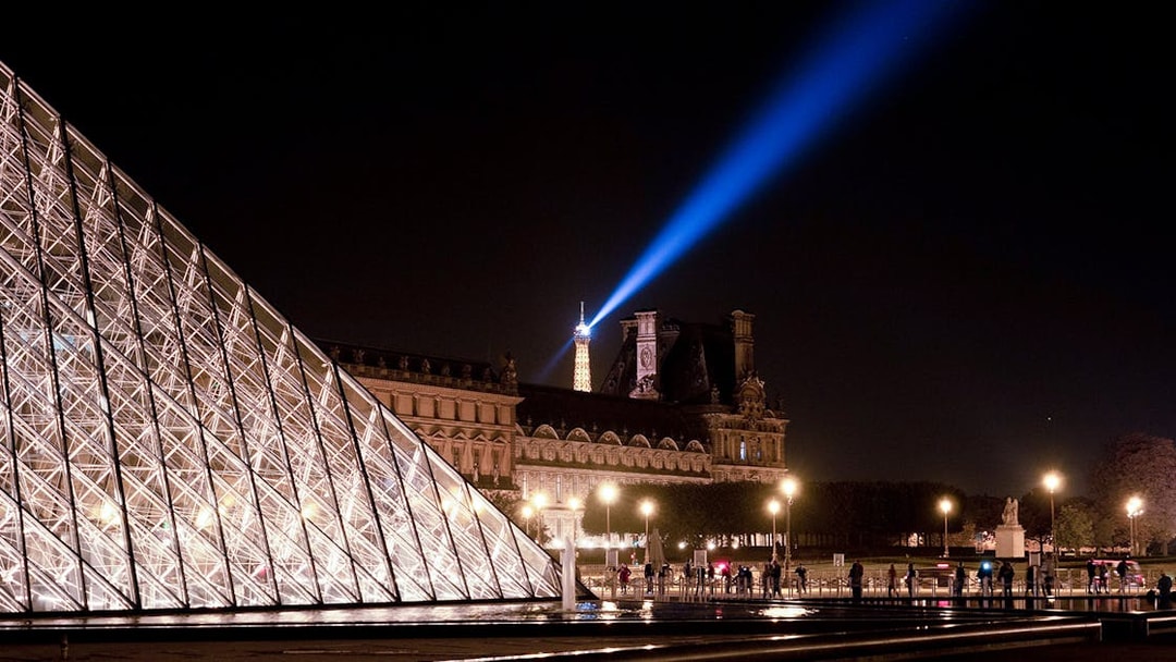 Louvre Pyramid illuminated at night with Eiffel Tower spotlight in Paris.