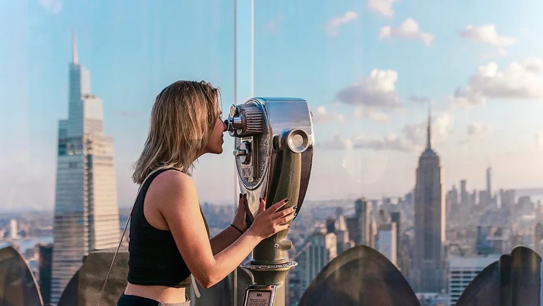 Woman using binoculars at Top of the Rock Observation Deck with Empire State Building view.