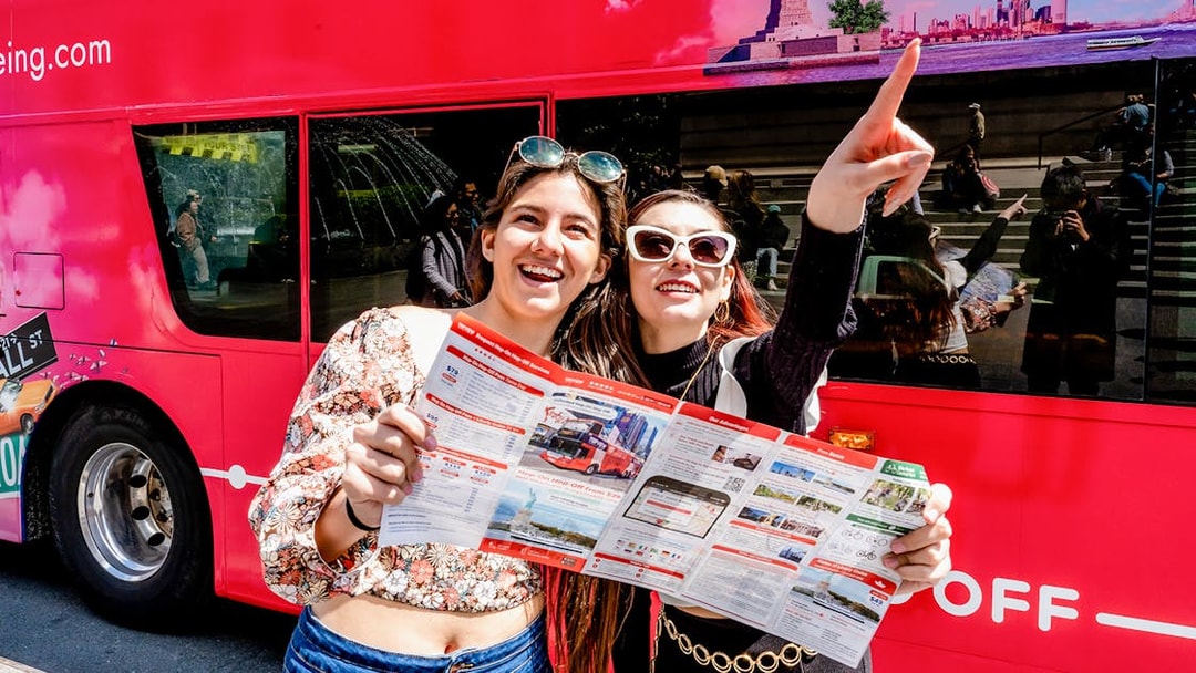 Tourists with map in front of red hop-on hop-off bus, pointing at city sights.
