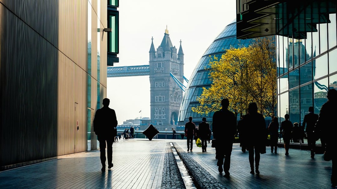 Silhouettes walking towards Tower Bridge, London, with modern buildings and trees.
