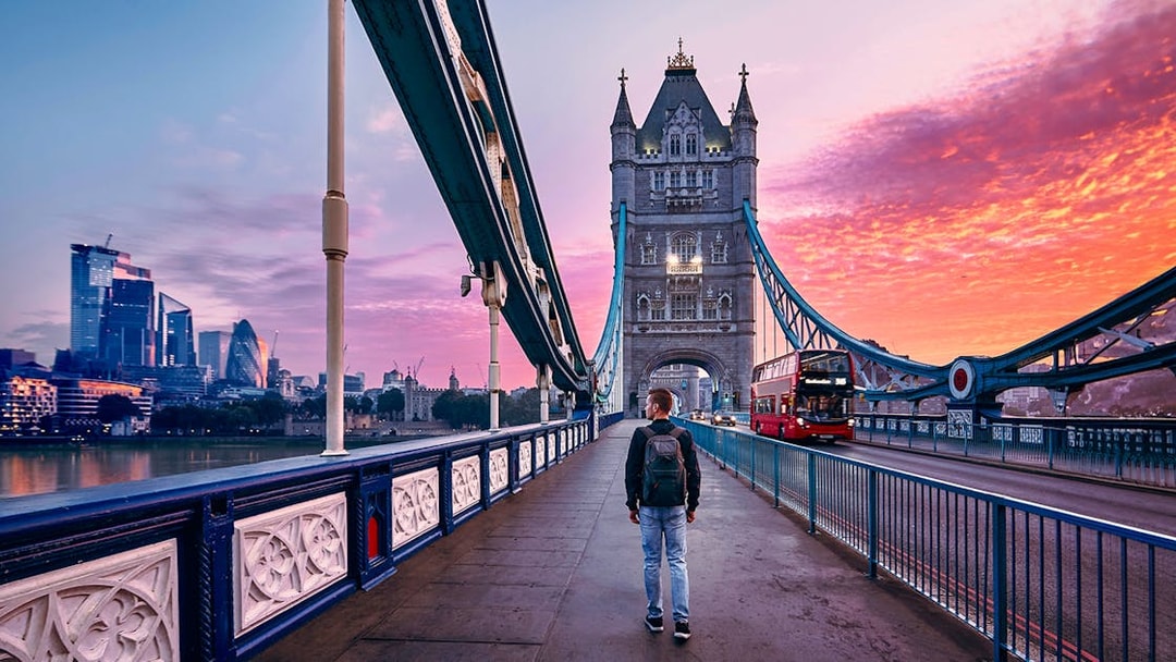 Person walking on Tower Bridge in London at sunset with a red bus passing by.
