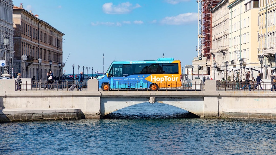 Hop-on hop-off bus crossing bridge in Trieste with historic buildings and Adriatic Sea in view.